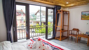 A hotel room with a bed, a wooden chair, a small table, and a balcony with a view of buildings and greenery.