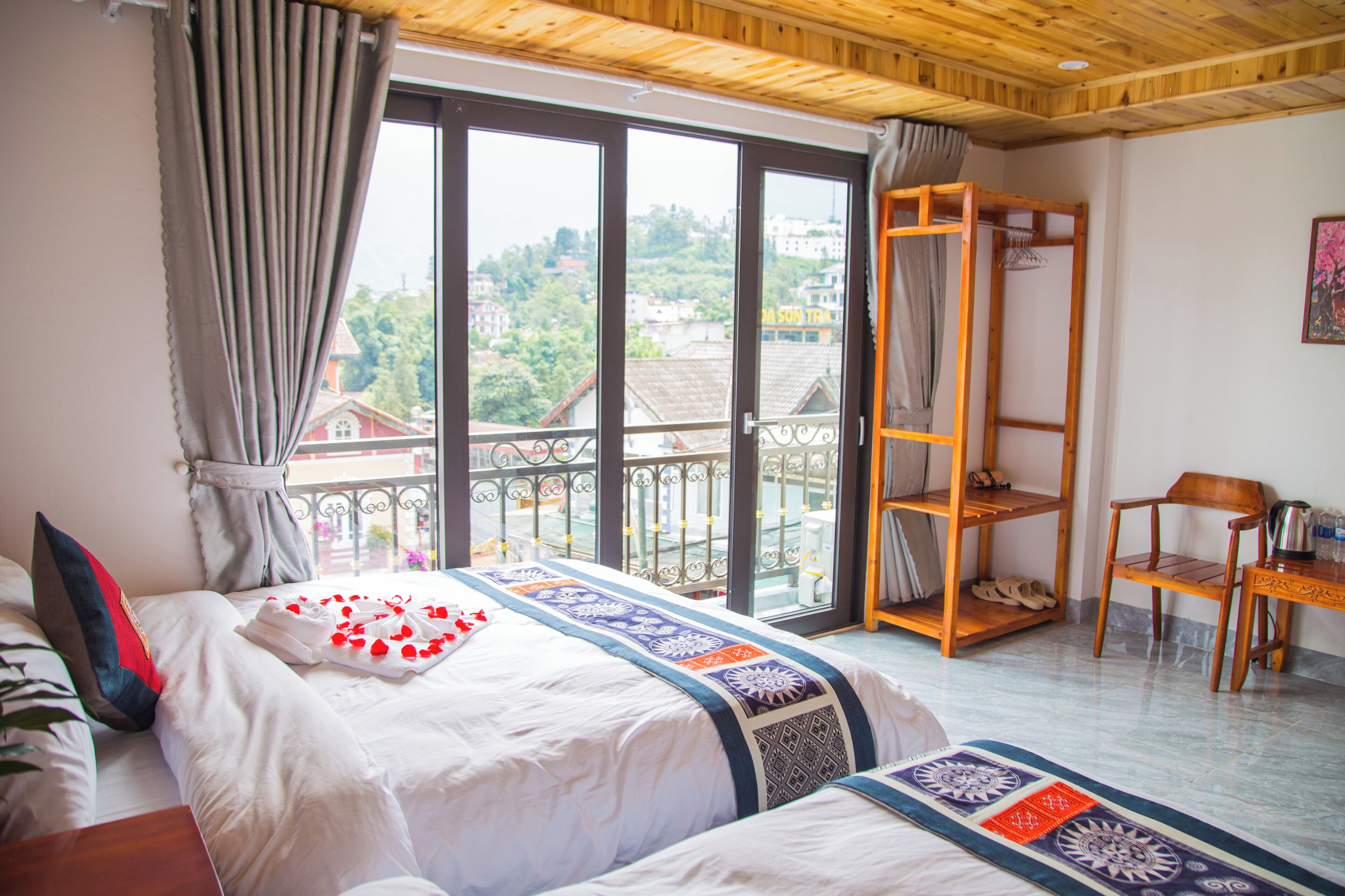 A hotel room with a bed, a wooden chair, a shelf, and a view of the city through large windows.