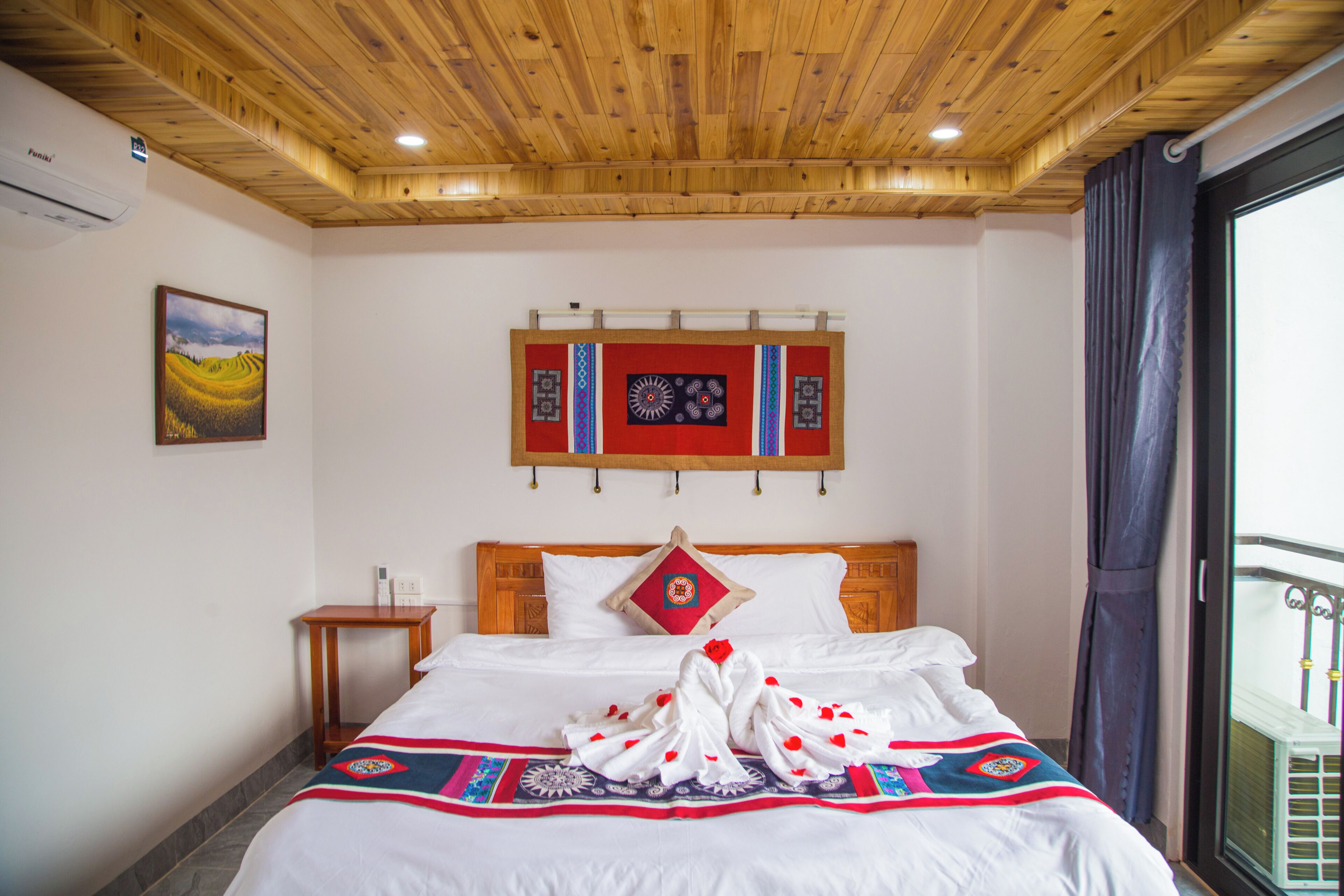 A hotel room with a wooden ceiling, a bed with a patterned bedspread, a wall-mounted artwork, and a window with curtains.