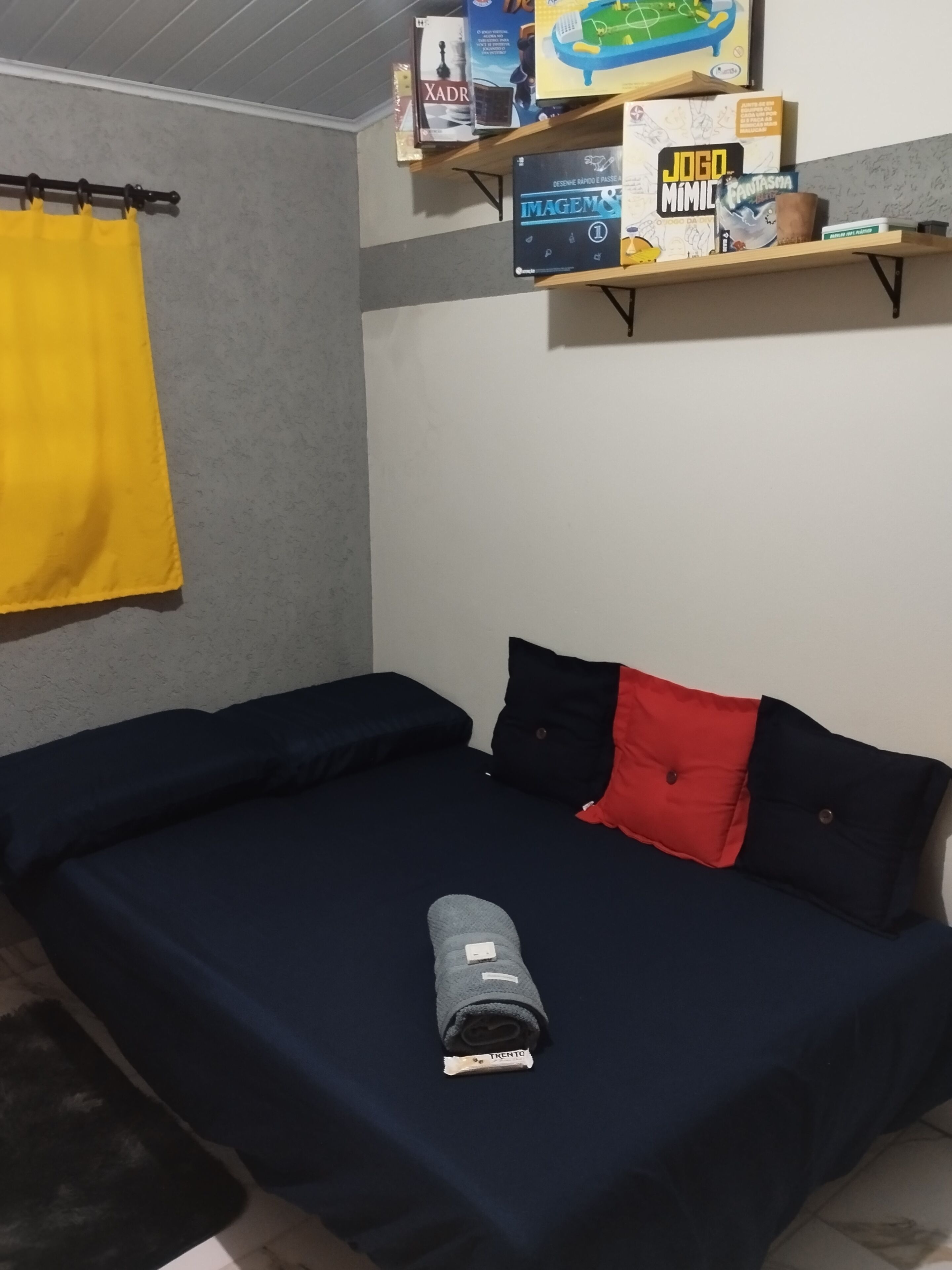 A room with a bed, a shelf with board games, and a yellow curtain.