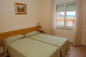 Una habitación con dos camas, una mesita de noche con lámpara, una ventana con cortinas e imagen en el muro.