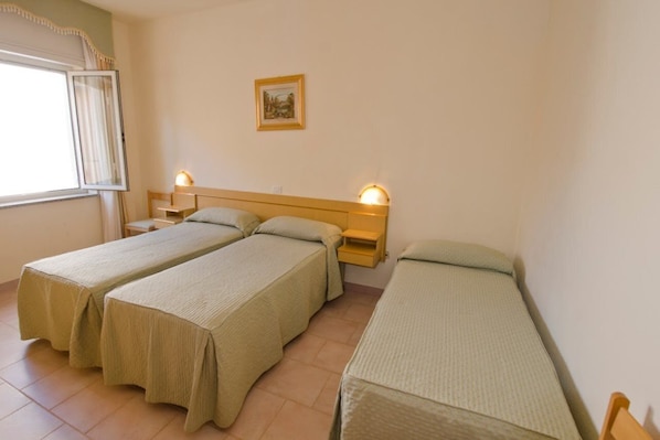 Una habitación con tres camas individuales, una mesita de noche con lámpara, un cuadro en la pared y una ventana con cortinas.