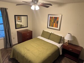 Une chambre à coucher comprenant un lit, une table de chevet, une commode, un ventilateur de plafond, une fenêtre avec des rideaux et deux tableaux encadrés accrochés au mur.