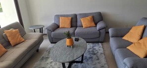 Ein Wohnzimmer mit grauem Sofagarnitur, zwei kleinen Beistelltischen und einem runden Couchtisch mit einer Pflanze.