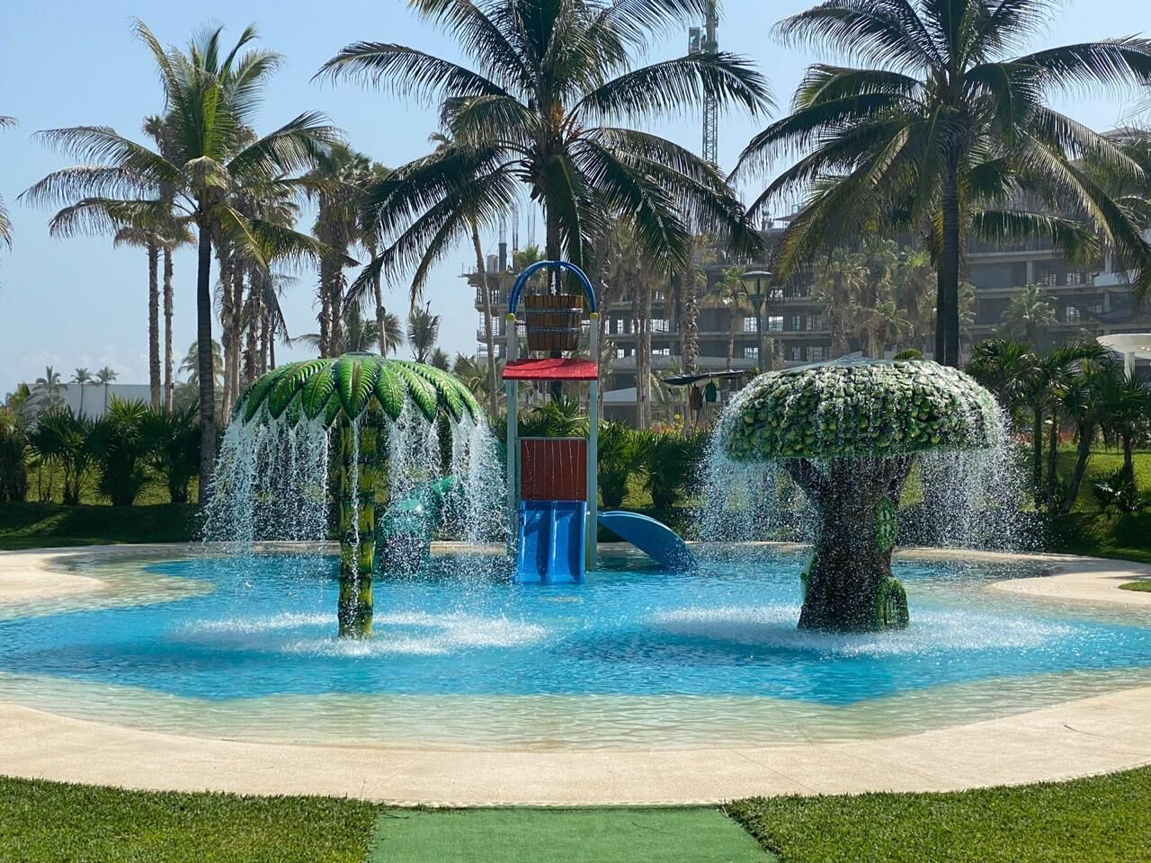 A water feature with a central fountain and two tree-shaped structures, surrounded by a pool and palm trees.