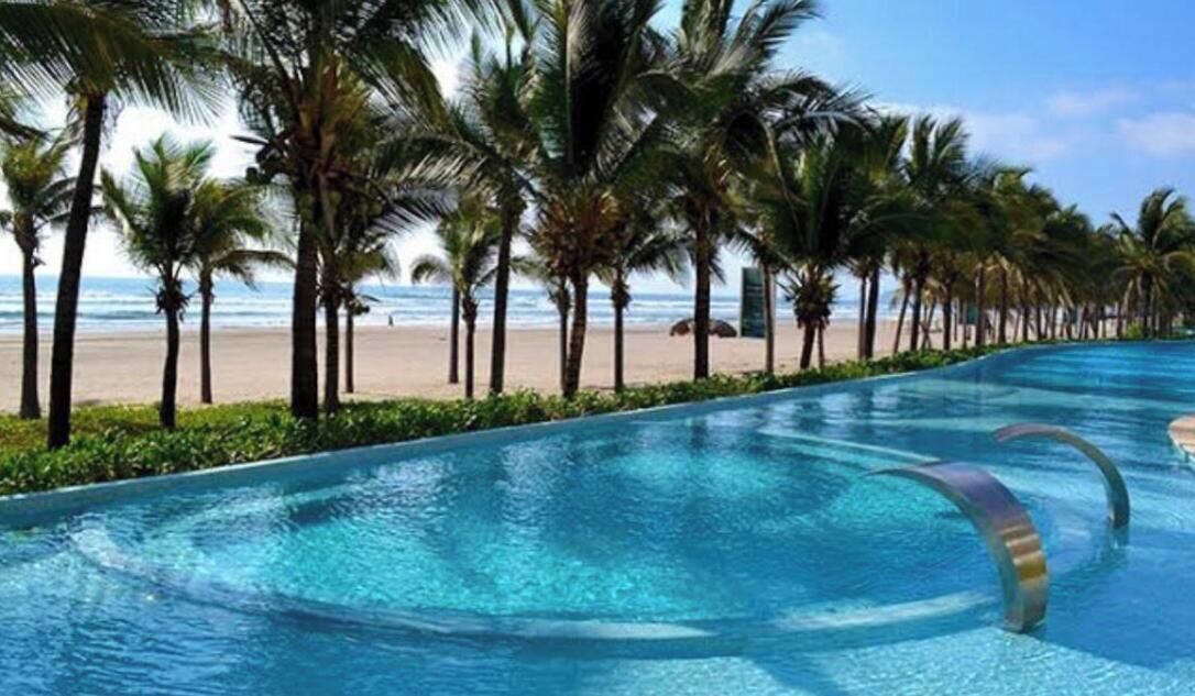Une piscine à l’eau d’un bleu cristallin, entourée de palmiers et avec une plage en arrière-plan.