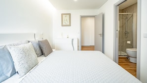 A bedroom with a bed, bedside tables, a framed artwork, and a bathroom with a shower and toilet. - Casas Da Alegria (Coimbra)
