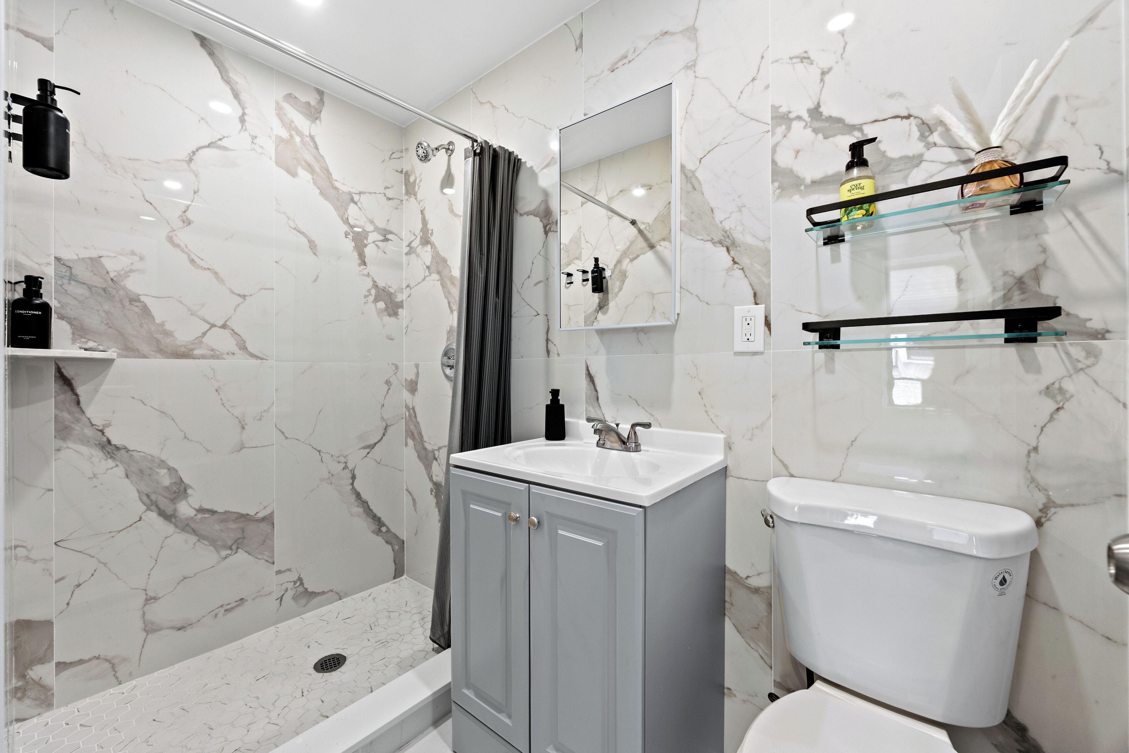 Een moderne badkamer met een marmeren spatwand, een grijze wastafelmeubel, een wit toilet en een glazen plank met toiletartikelen.