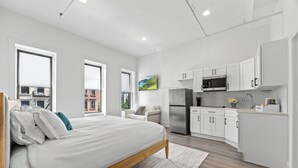 A modern bedroom with a large bed, a sofa, and a kitchen area with white cabinets and appliances.