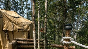 Señal de madera que indica 'Spruce Hollow Tent 1' en un área boscosa, con una tienda de campaña y una puerta de cuerda.
