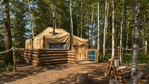 A rustic tent with a chimney, surrounded by a wooden fence and a stack of logs.