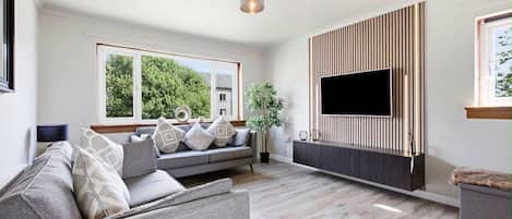 A modern living room with a grey sofa, a flat-screen TV mounted on a wooden panel, and a large window offering a view of greenery.