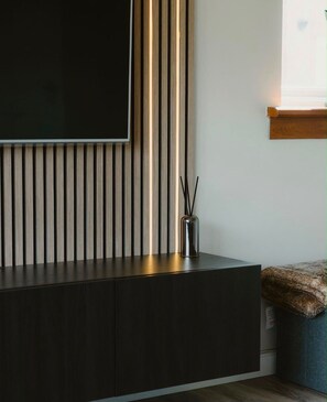Ein modernes Wohnzimmer mit einem dunklen Holzschrank, einem flachbildfernseher an einer Wand mit vertikalen Holzbalken und einer kleinen Vase mit Zweigen.