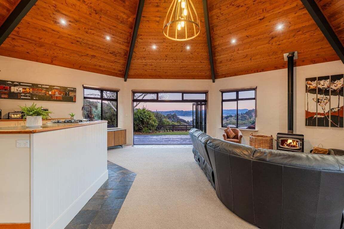 Ein modernes Interieur mit Holzbalkendecke, einer Küchenzeile, einer Sitzecke mit Kamin und großen Fenstern mit Blick ins Freie.