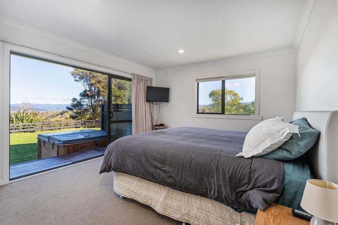 A bedroom with a large bed, a television, and a window with a view of a garden and hills.