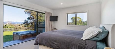 A bedroom with a large bed, a television, and a window with a view of a garden and hills.