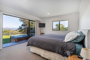 A bedroom with a large bed, a television, and a window with a view of a garden and hills. - La Casa Escondida (Whitianga)
