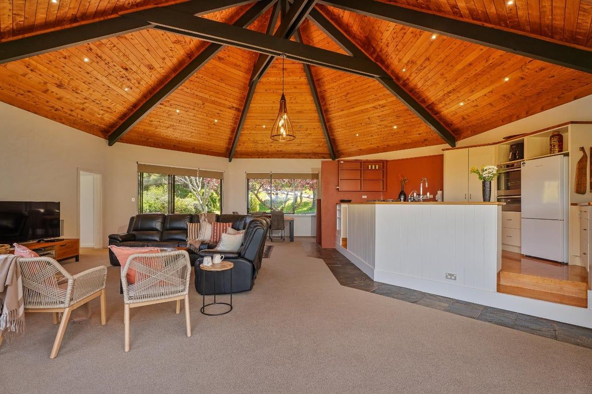 A spacious living area with a high ceiling, a black leather sofa, wicker chairs, and a kitchen area with white cabinets and a countertop.