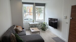 Un salón moderno con un sofá gris, una mesa de centro, un televisor montado en la pared y un ventanal con vistas a la calle.