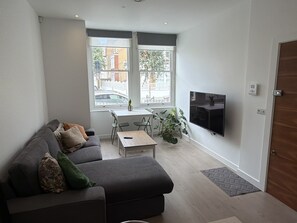 Ein modernes Wohnzimmer mit grauem Sofa, Couchtisch, Wandfernseher und Fenster mit Blick auf die Straße.