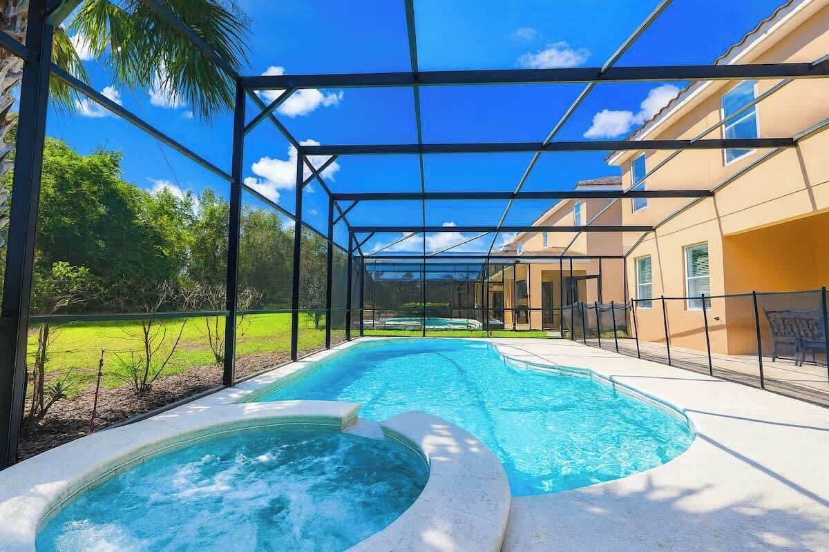Une piscine avec un jacuzzi, entourée d’une zone couverte dotée d’une structure métallique et de murs en verre, et un bâtiment en arrière-plan.