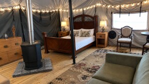 A tented accommodation with a bed, bedside lamps, a sofa, a table, and a wood-burning stove.