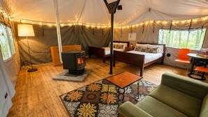 A tented accommodation with a central wood-burning stove, two beds, a sofa, a table, and string lights.