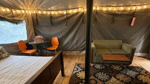 A tent interior with a bed, two orange chairs, a green sofa, a small table, and a patterned rug.