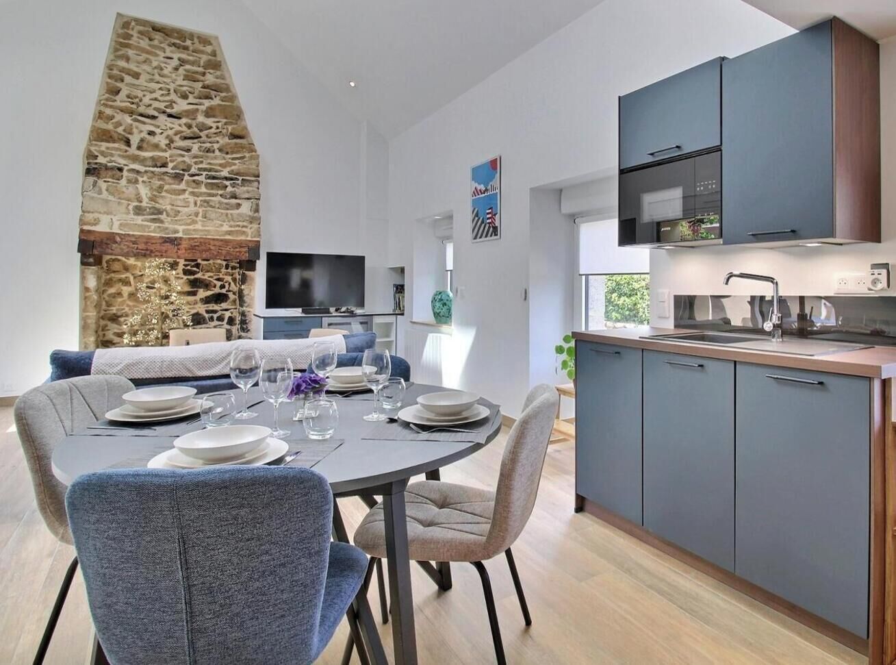 Une cuisine moderne avec des meubles gris, une cheminée en pierre et un coin repas avec une table ronde dressée pour quatre personnes.