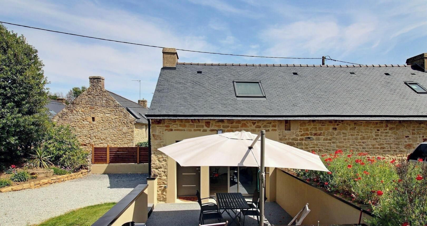 Un bâtiment en pierre avec un toit en pente, une terrasse dotée de mobilier d’extérieur et un jardin agrémenté de fleurs rouges.