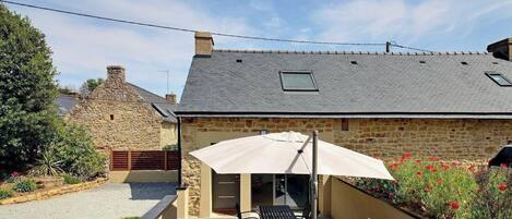 Un bâtiment en pierre avec un toit en pente, une terrasse dotée de mobilier d’extérieur et un jardin agrémenté de fleurs rouges.