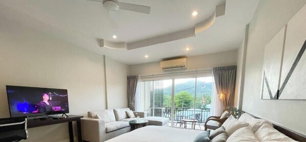 A hotel room with a bed, a TV, a balcony with a table and chairs, and a view of greenery outside.