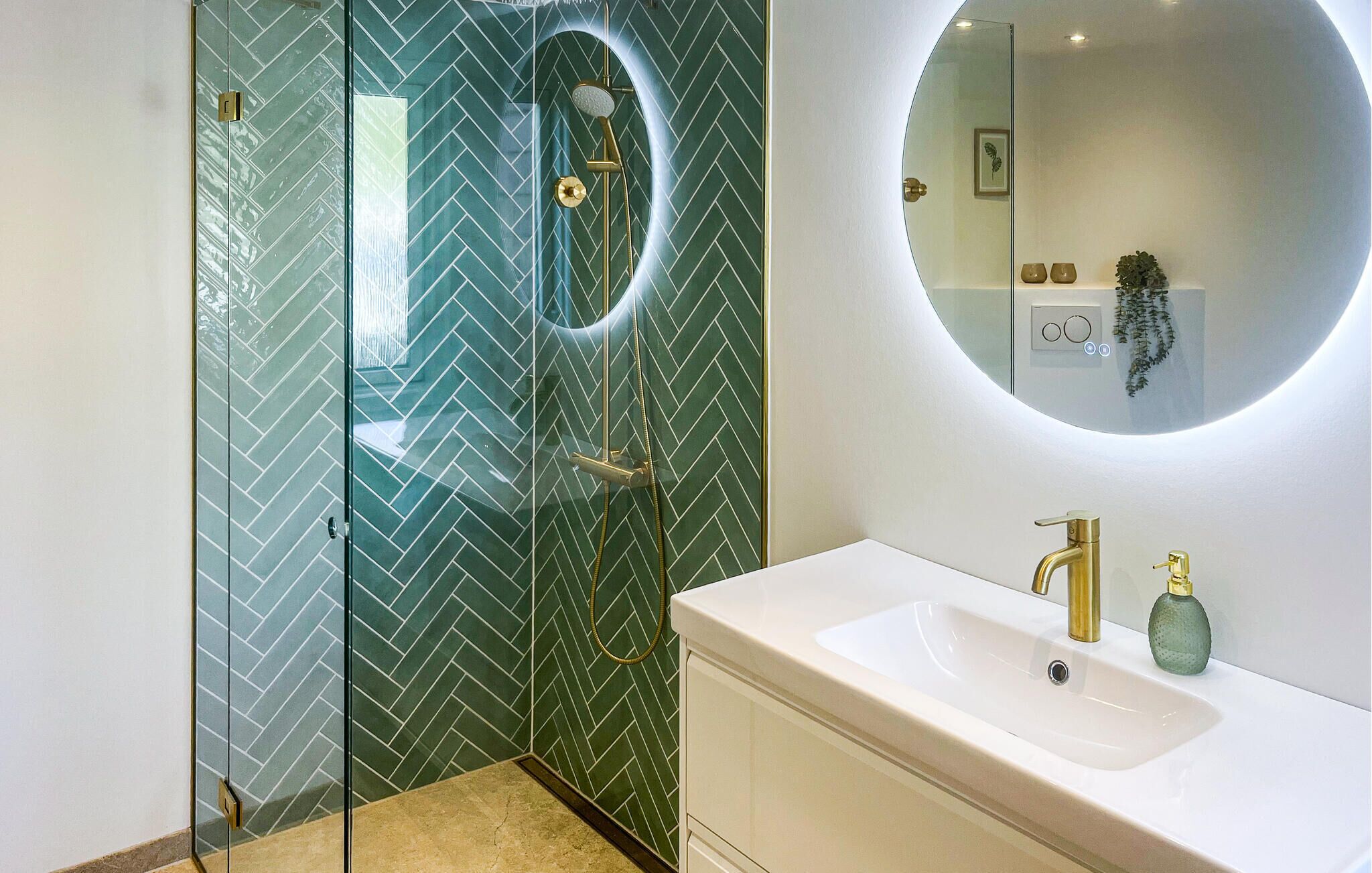 Une salle de bain moderne avec une douche à parois de verre, un lavabo blanc doté de robinetterie dorée et un miroir rond.