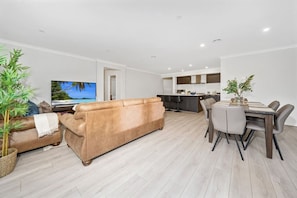 A modern living room with a sofa, a dining area with a table and chairs, a kitchen in the background, and a television on the left. - Comfy Premium 4BR Near Airport, Cafes & Amenities (Mickleham)