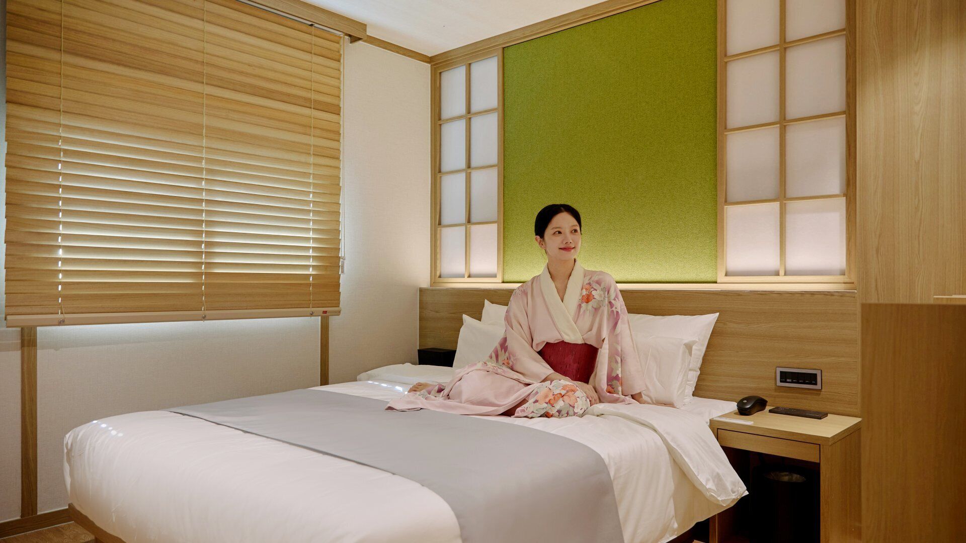 A hotel room with a bed, a nightstand, a wall-mounted control panel, and a window with blinds.