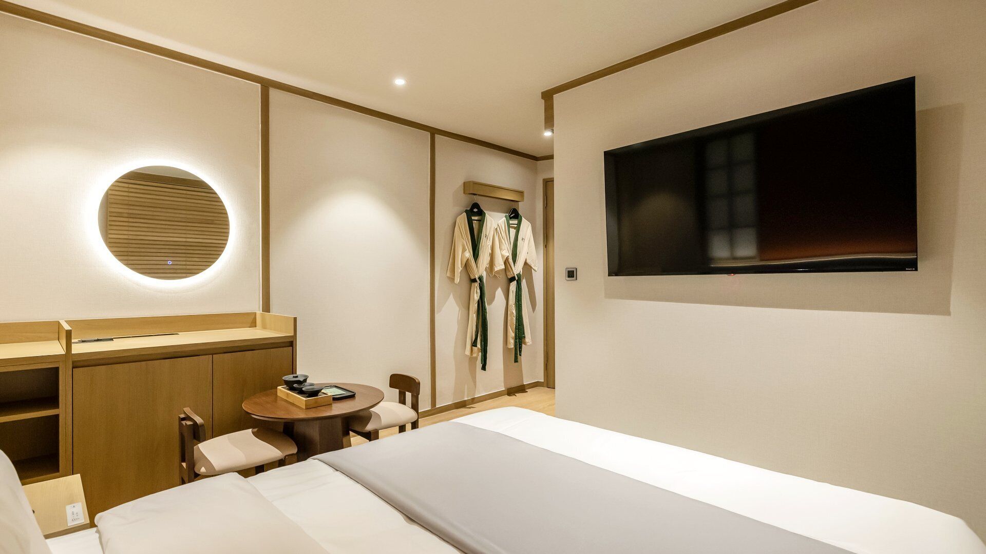 Habitación de hotel con cama, espejo redondo, televisión montada en la pared, una pequeña mesa con una silla y perchero con batas.