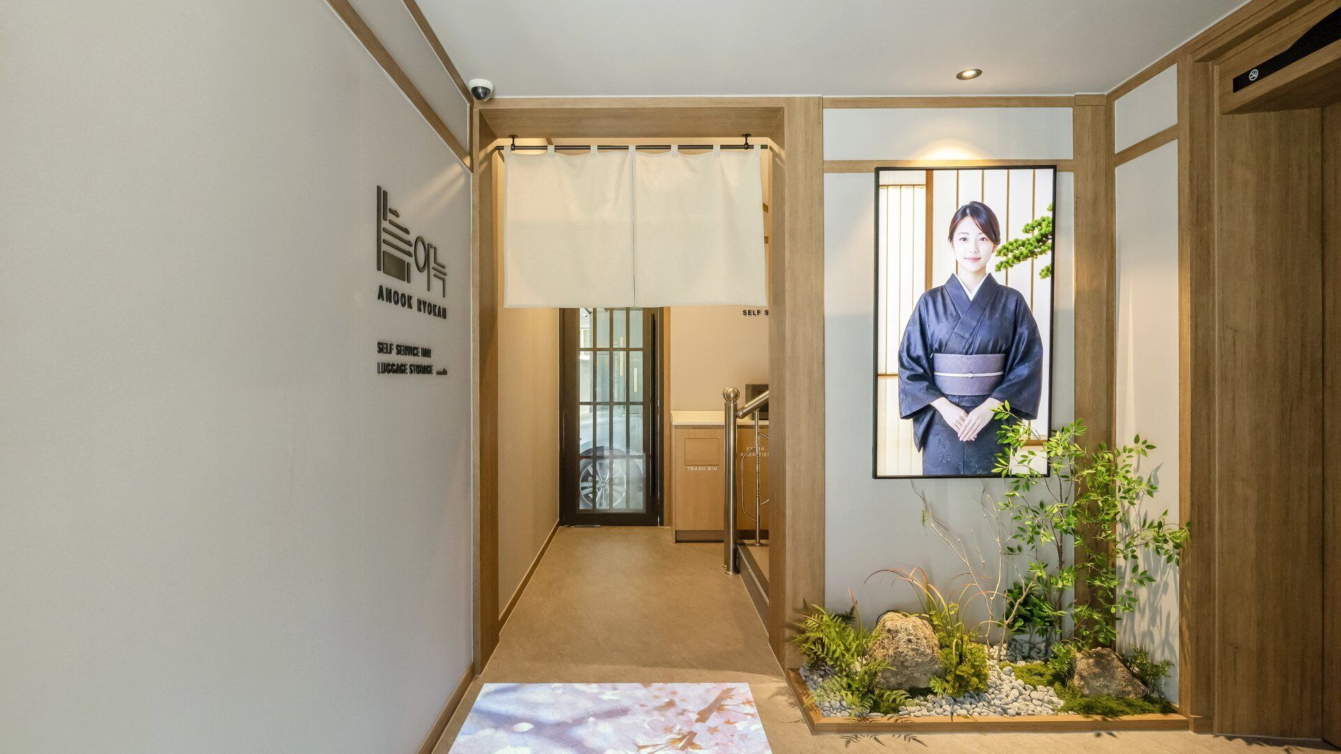 Un pasillo con estética tradicional japonesa, que incluye un tatami, un cuadro enmarcado de un kimono y un pequeño jardín interior.