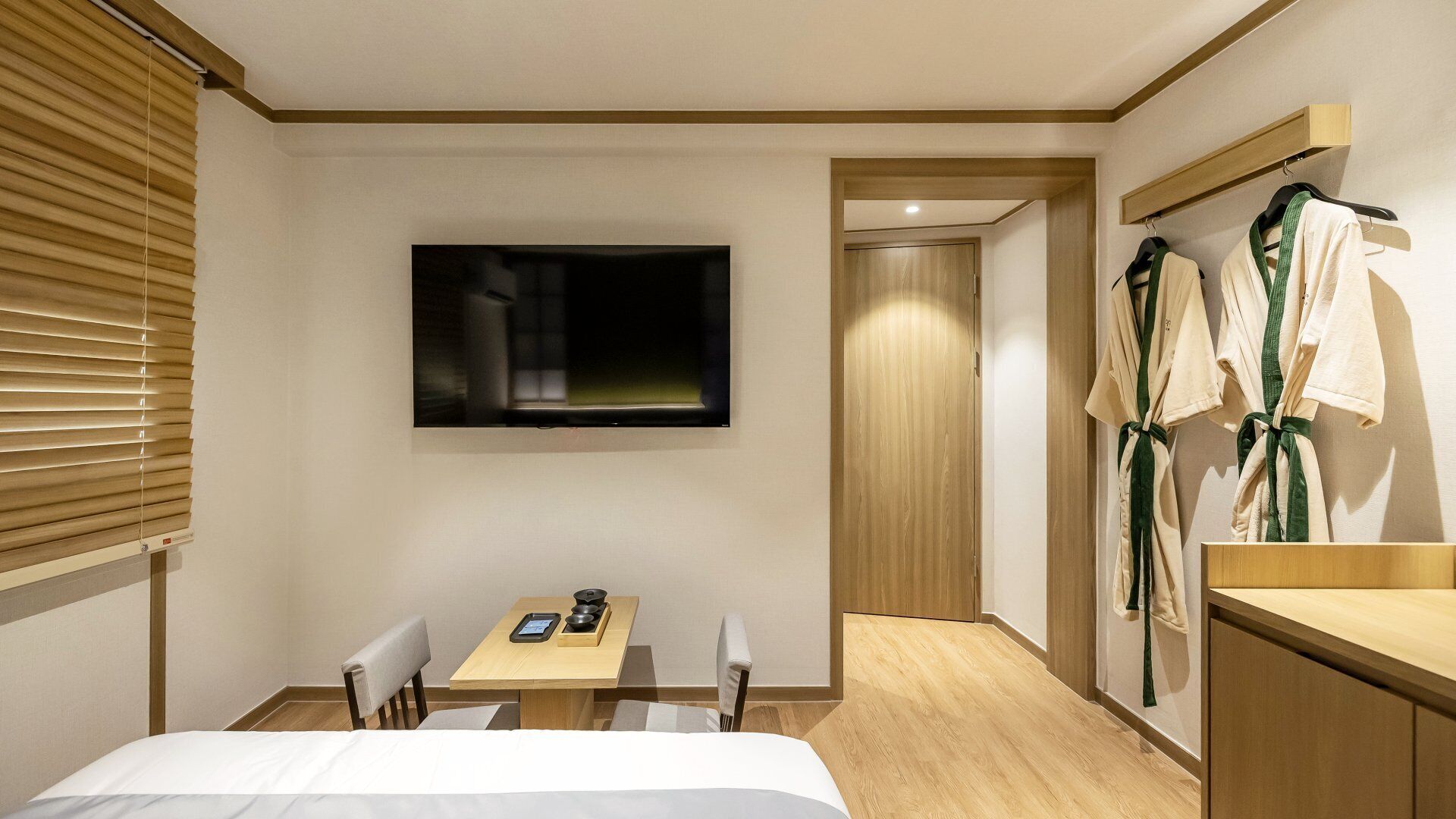 A hotel room with a bed, a table with a tablet and a remote, a TV on the wall, and a robe rack with robes.