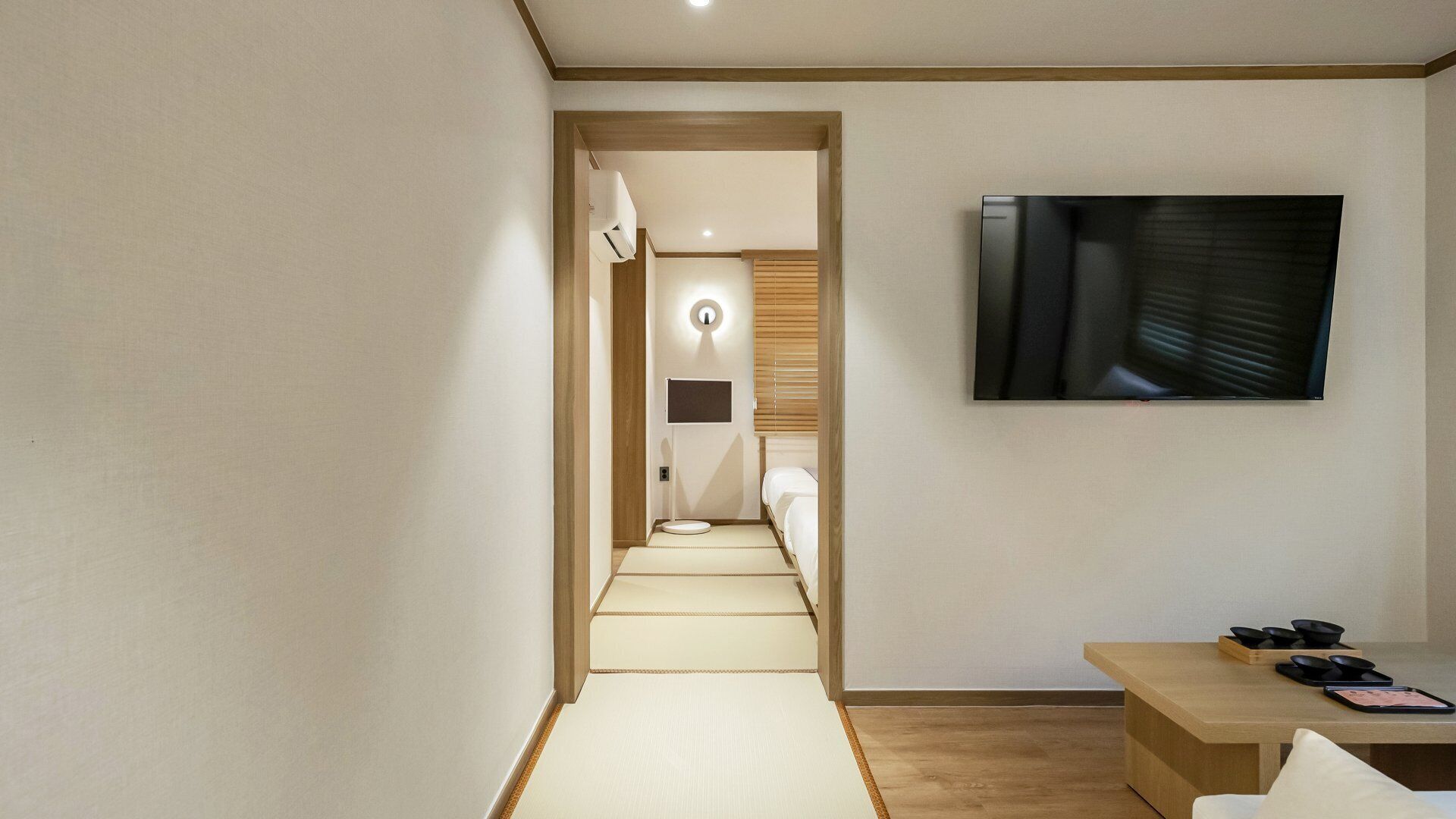 A hotel room with a television mounted on the wall, a small table with a tray of bowls, and a tatami mat leading to a bathroom.
