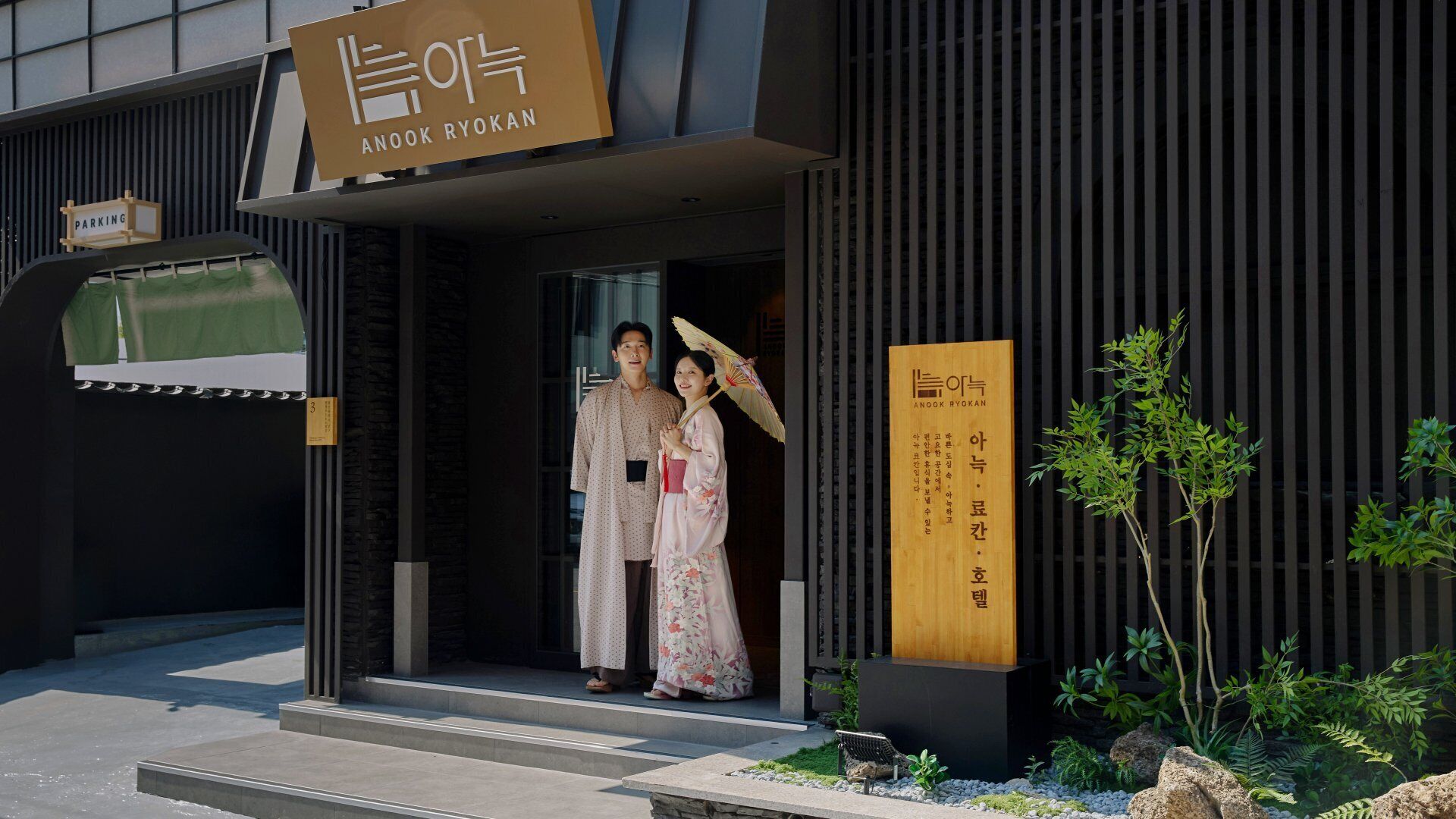 A building entrance with a sign reading 'ANOOK RYOKAN' and a traditional Korean couple standing at the doorway.