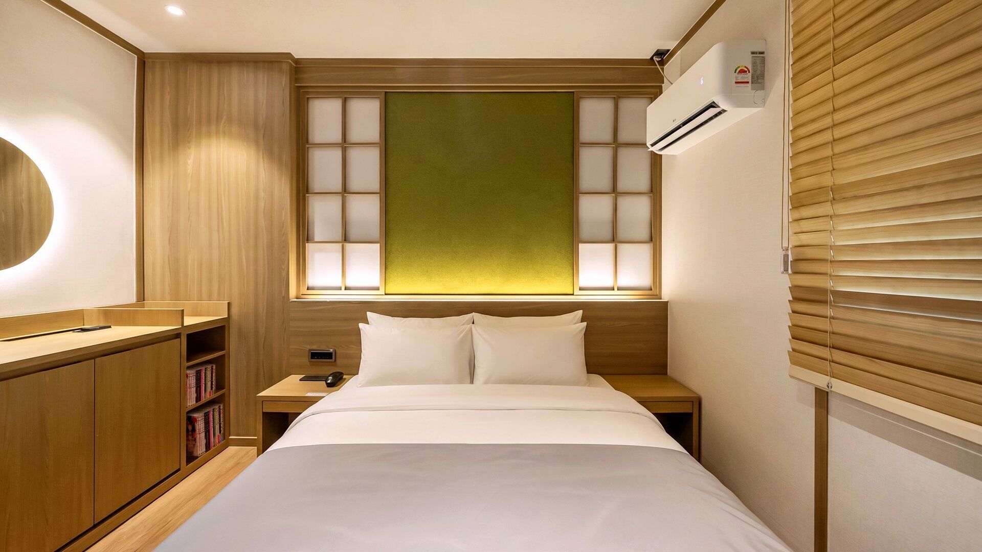 A hotel room with a bed, bedside tables, a nightstand, a bookshelf, and a window with blinds.