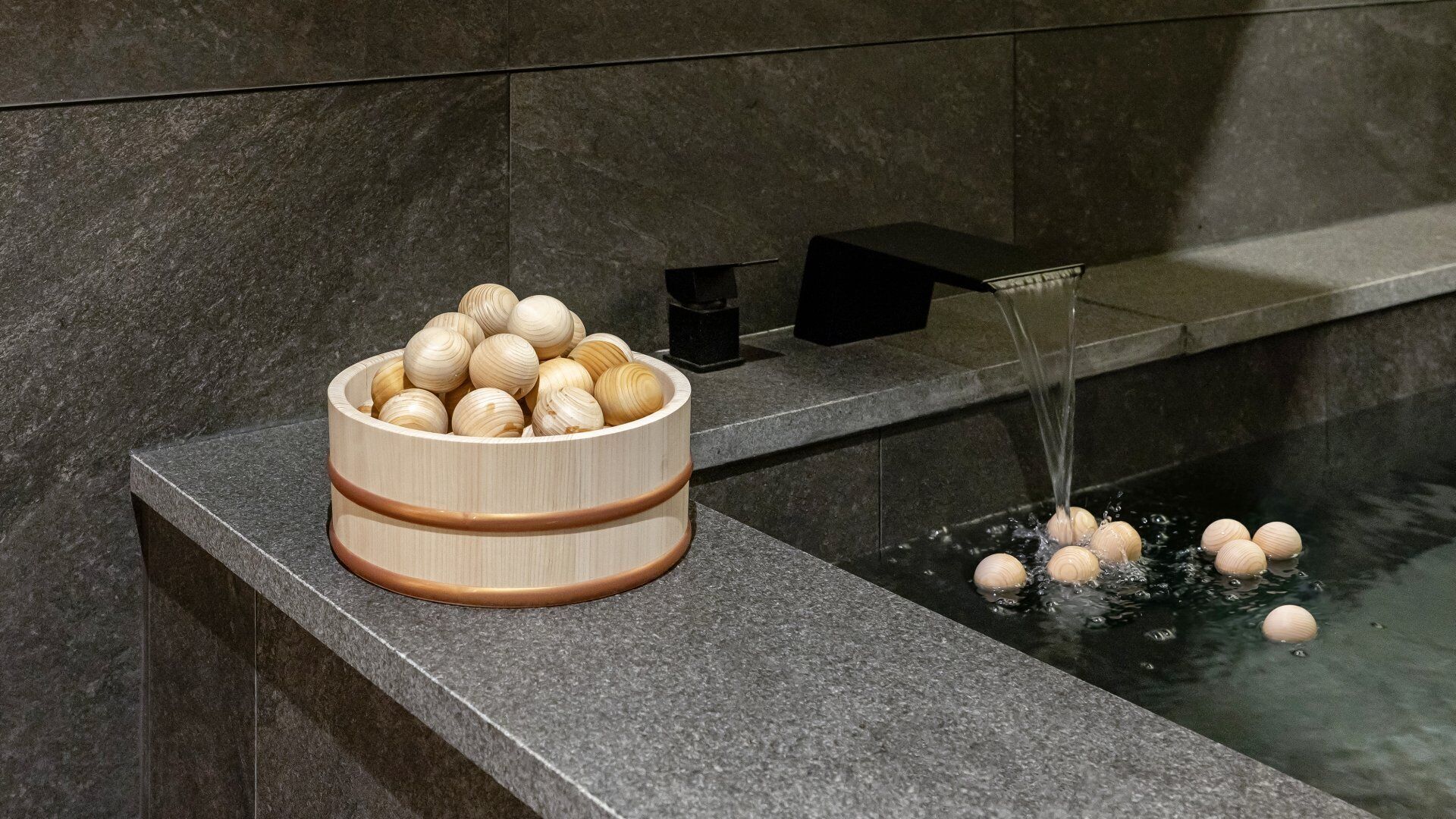 A wooden bowl filled with round objects on a stone ledge next to a water feature.