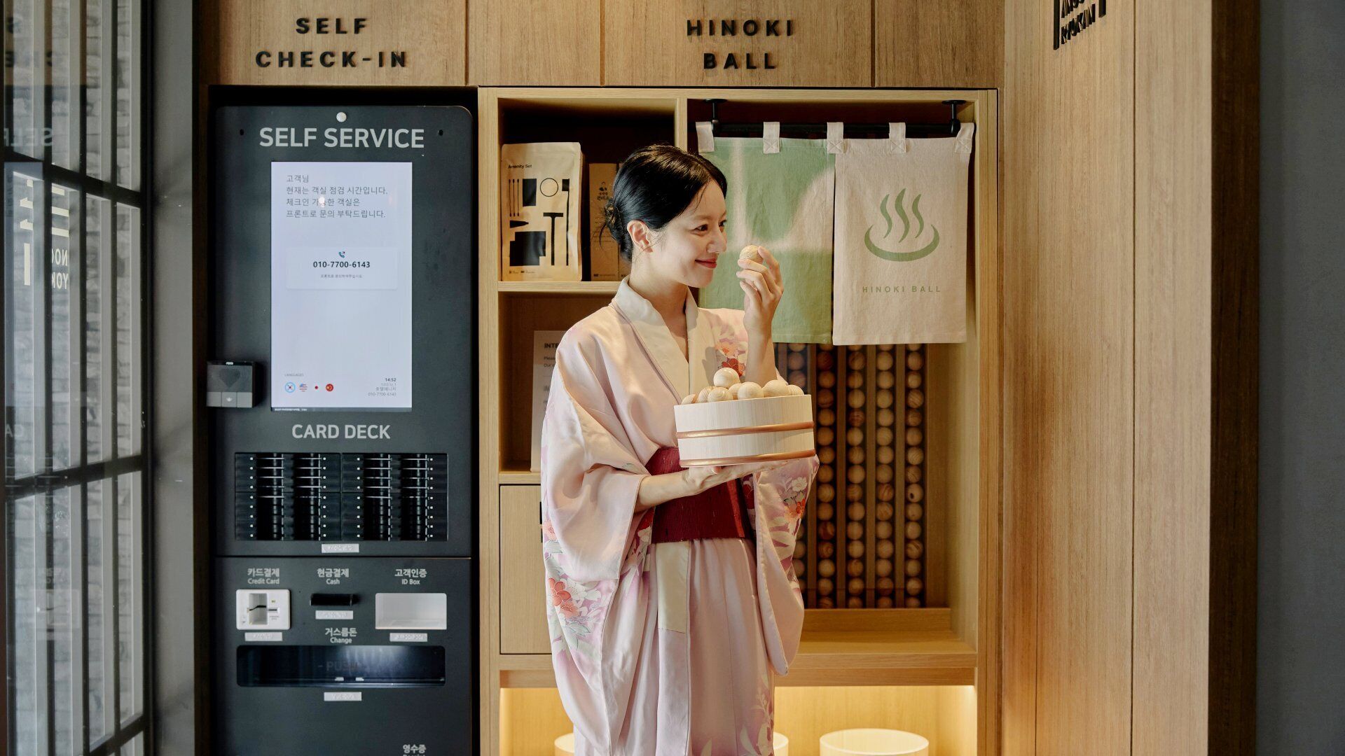 A self-service kiosk with card deck and ID box, a Hinoki Ball display, and a person in traditional attire holding a tray.