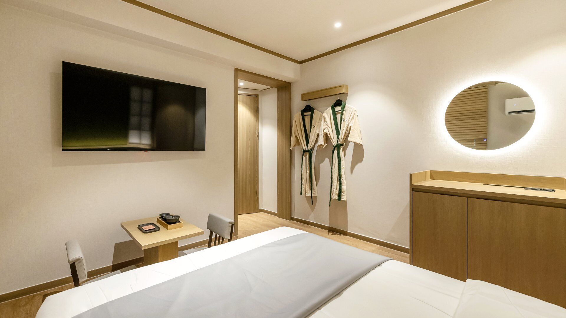 A hotel room with a bed, a desk with a chair, a TV mounted on the wall, and a mirror with a circular frame.