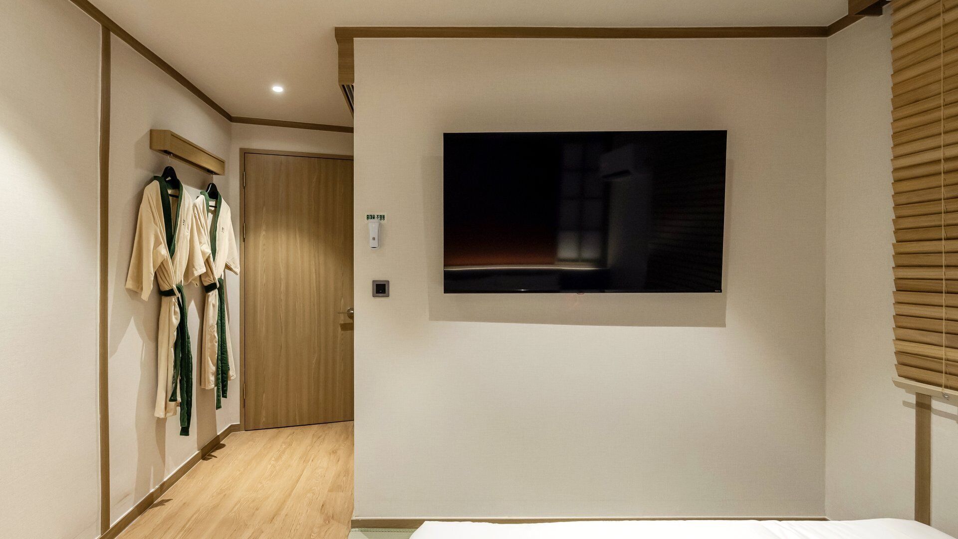 A hotel room with a bed, a wall-mounted TV, a wooden door, and a coat rack with robes.