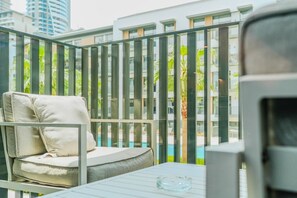 Ein Balkon mit weiß gepolstertem Stuhl, einem Glastisch und Blick auf einen Swimmingpool sowie moderne Gebäude.