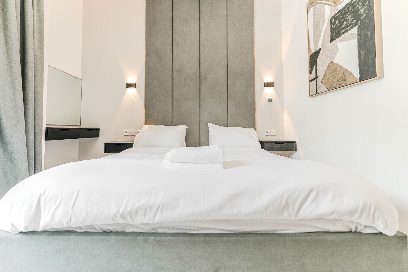 A modern hotel room with a large bed, two bedside tables, a headboard, and a wall-mounted artwork.