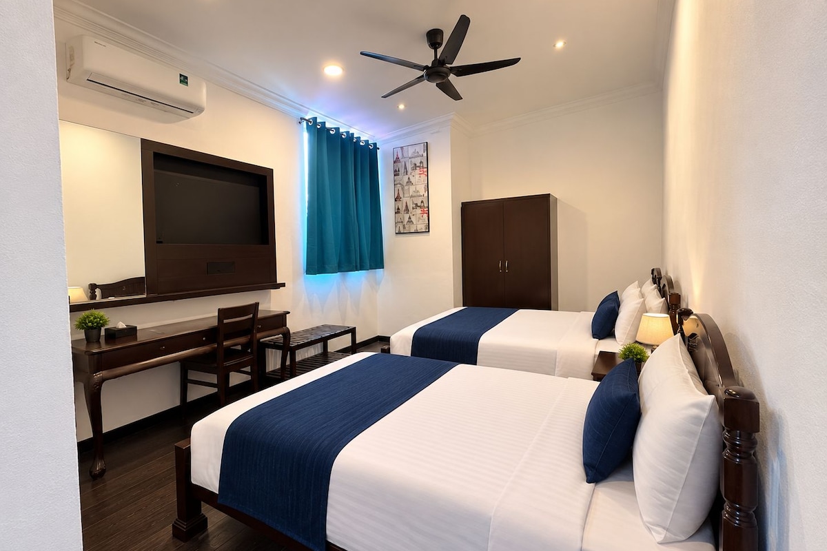 A hotel room with two beds, a desk, a chair, an air conditioning unit, a ceiling fan, and a window with curtains.