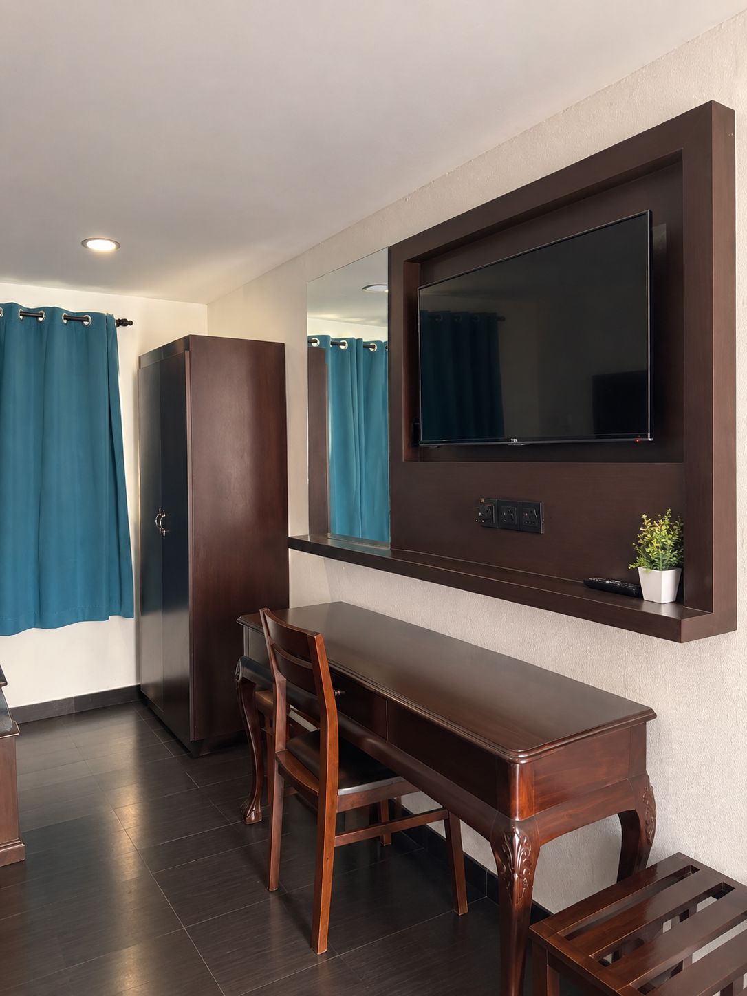 A hotel room with a wooden dining table, chairs, a TV mounted on the wall, a wardrobe, and a shower area with curtains.
