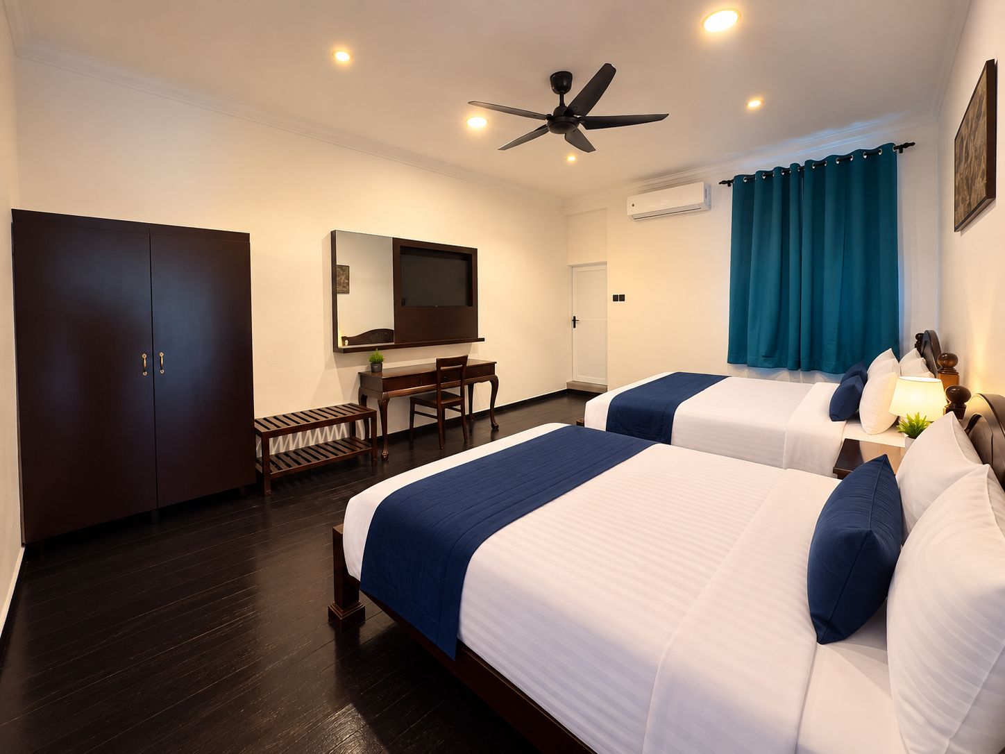 A hotel room with two beds, a desk, a chair, a television, a ceiling fan, and a wardrobe.